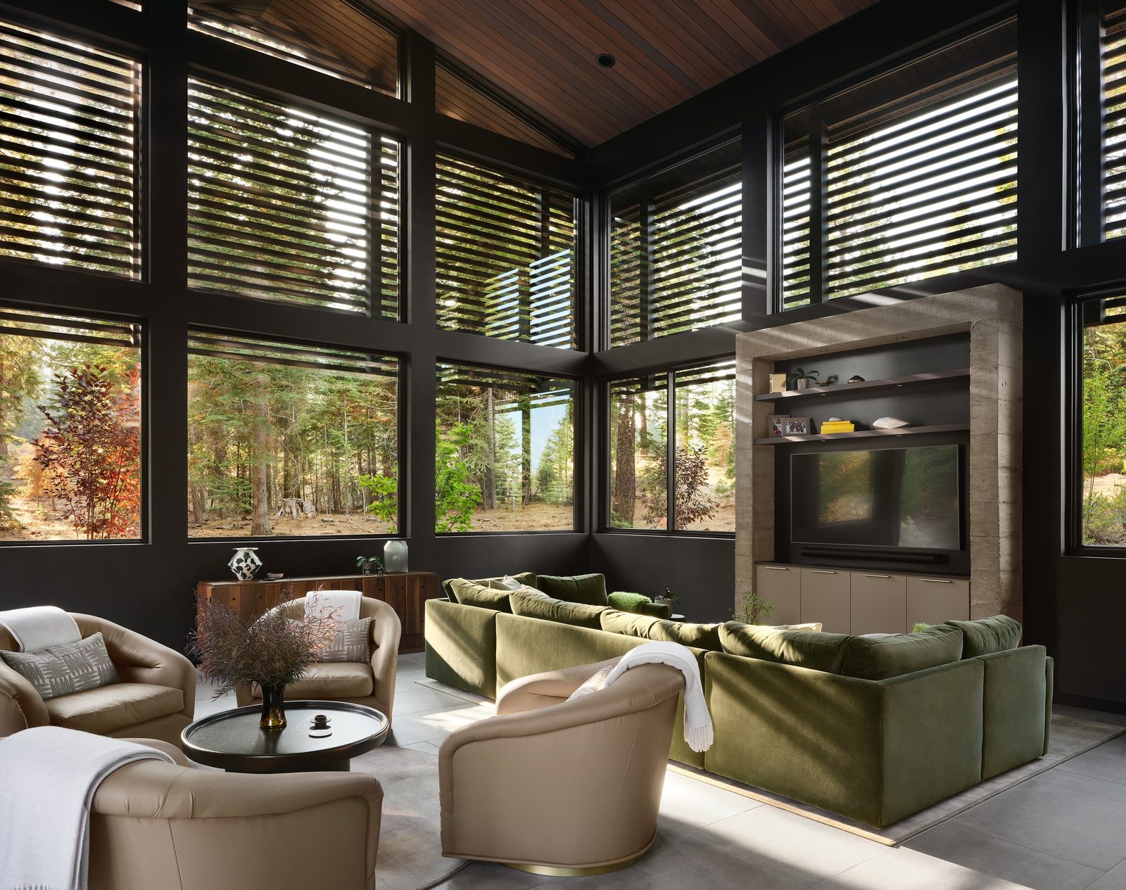 Great room by Hills & Grant Interior Design with an olive velvet sectional, stone media wall and glass walls onto pines